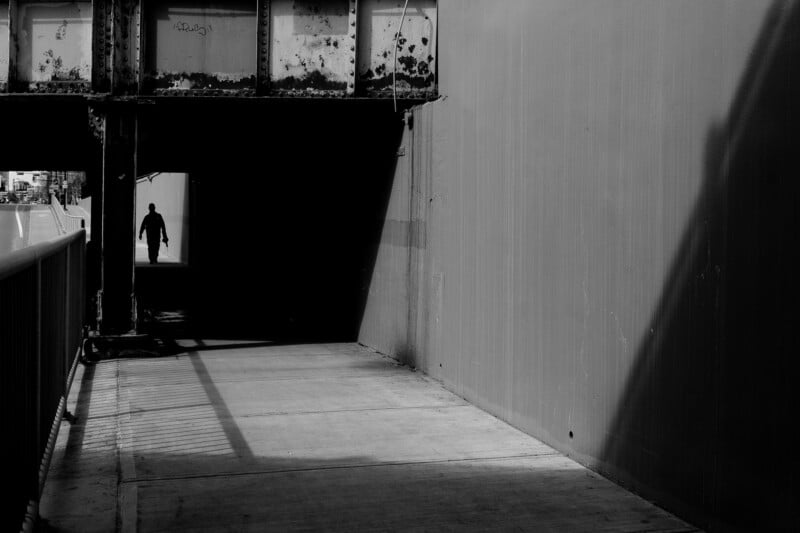 A person walks through a shadowy underpass beneath a rusted bridge, silhouetted against a bright opening at the far end. The scene features strong contrasts between light and dark areas.