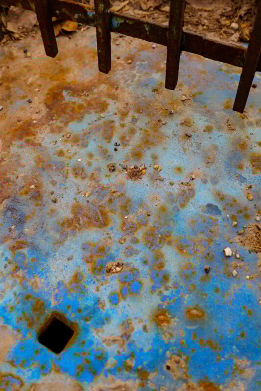 Rusty blue metal surface with scattered brown spots, small pebbles, and a single square hole in the lower left. Part of a weathered metal fence appears at the top. Some dry leaves and dirt are visible around the edges.