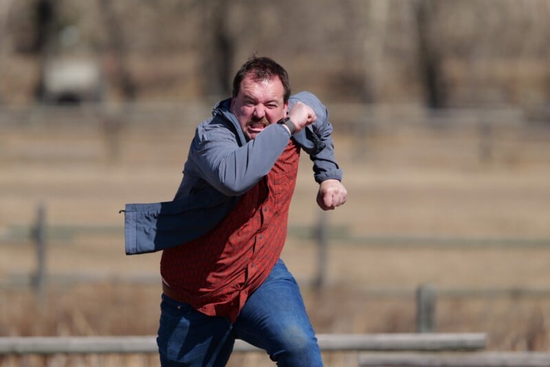 A man outdoors in a red shirt and gray jacket is captured mid-motion, running with his arm bent in front of his face and an intense expression. The background is blurred, with wooden fences and dry grass visible.