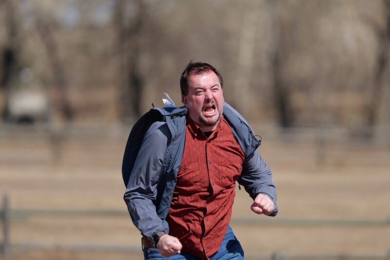 A man with a backpack runs outdoors, clenching his fists and yelling with an intense, angry expression. The background is blurred, showing bare trees and a field.