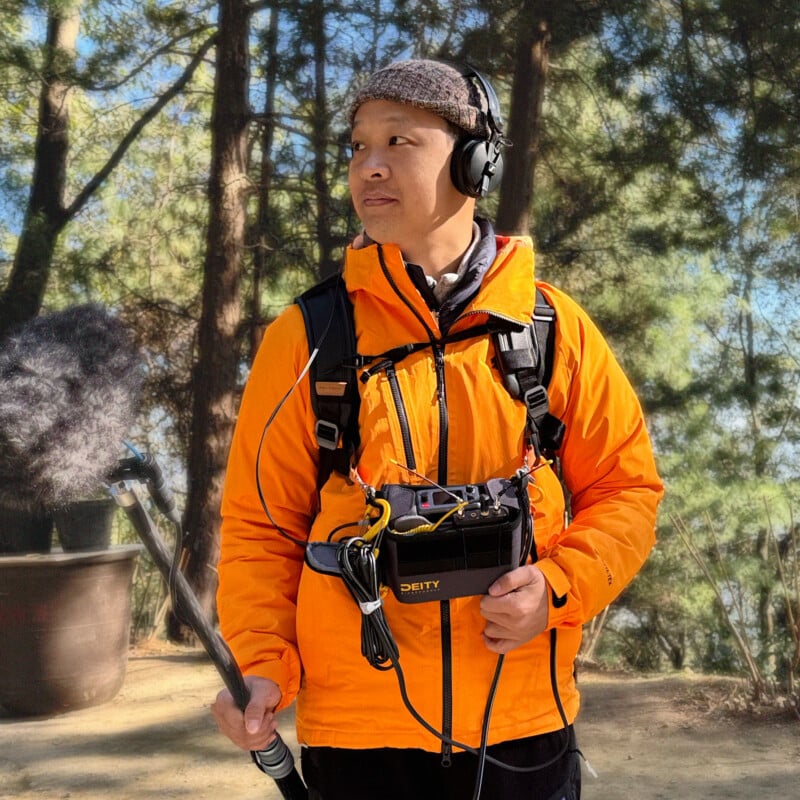 A person in an orange jacket and headphones holds a boom microphone and audio equipment while standing in a sunlit forest.