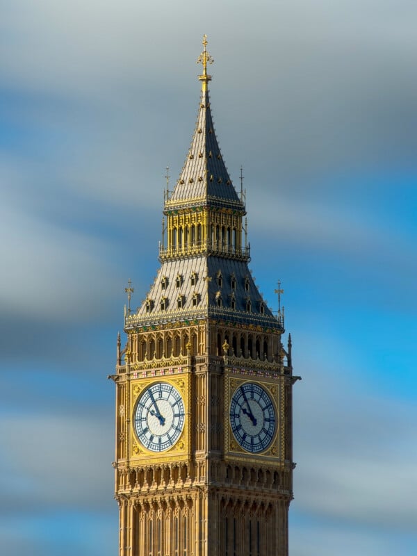 The image shows the clock tower known as Big Ben in London against a blue sky with some wispy clouds. The ornate details and clock faces are clearly visible.
