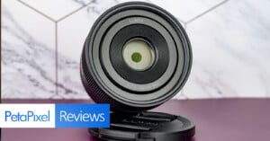 A Lumix S 1:2/40mm lens stands upright on a purple surface with its lens cap beside it. The background features a white and gray geometric pattern. “PetaPixel Reviews” is labeled in the bottom left corner.