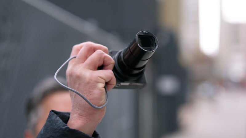 A person holds a digital camera with a large zoom lens, pointing it upwards. The background is blurred, showing an outdoor urban setting with buildings and a chain-link fence.