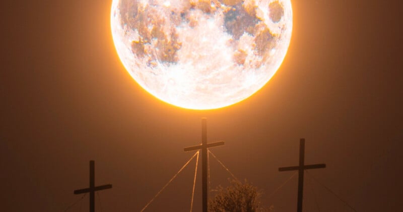 Una luna llena grande y brillante parece descansar sobre la cruz central, con otras dos cruces cercanas, recortadas contra el cielo nocturno. La escena crea una atmósfera poderosa y etérea.