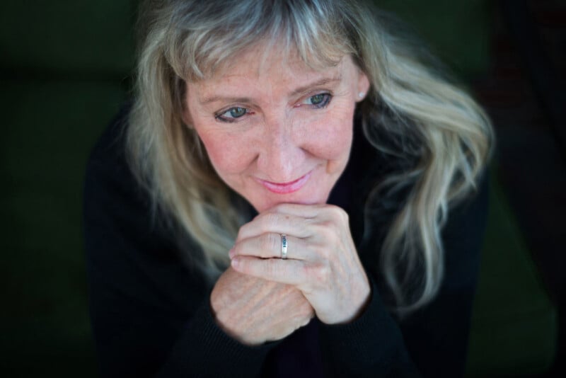 A middle-aged woman with long blond hair rests her chin on her hands, smiling softly and looking to the side. She wears a black top and a simple ring on her finger.