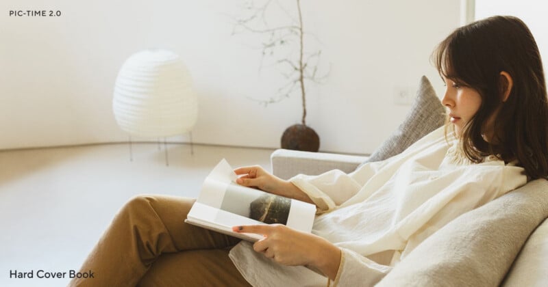 A woman sits on a light-colored sofa in a minimalist room, reading a hardcover book. There is a white paper lamp and a bare branch in a pot in the background. The atmosphere is calm and relaxed.