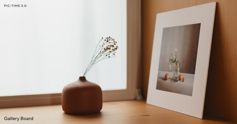 A small brown vase with dried flowers sits on a wooden surface next to a framed photo of a vase with flowers and fruit, placed by a softly lit window.