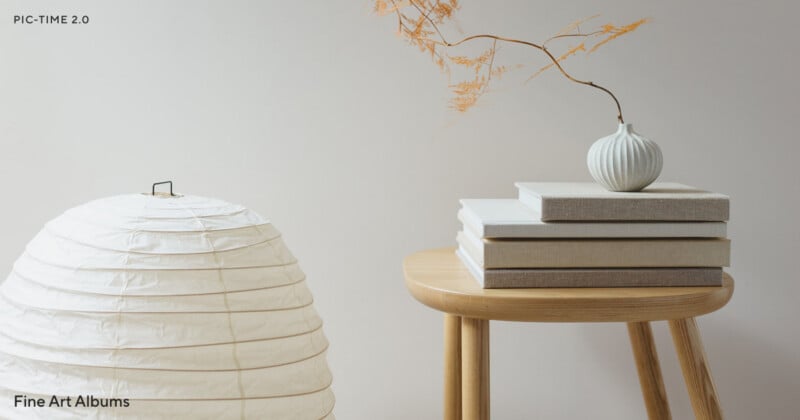 A minimalist scene with a round paper lantern, stacked neutral-colored photo albums on a wooden stool, and a small vase holding delicate dried branches against a plain light background.