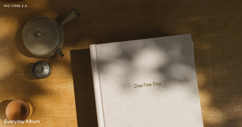 A linen photo album titled "One Fine Day" rests on a wooden table beside a teapot, teacup, and shadows of leaves. The text "PIC-TIME 2.0" and "Everyday Album" appear in the corners.