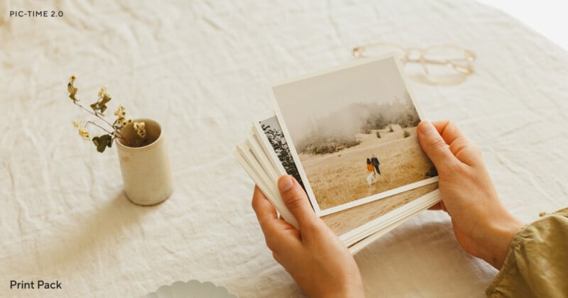 A person holds a stack of printed photos, with the top photo showing a couple walking in a field. The scene is set on a light fabric surface with a small vase of dried flowers and a pair of eyeglasses nearby.