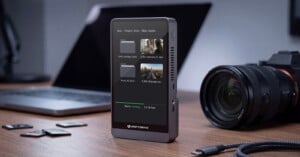 A portable external drive with a screen shows file folders and photo thumbnails, sitting on a desk near a laptop, a camera, SD cards, and a USB cable.