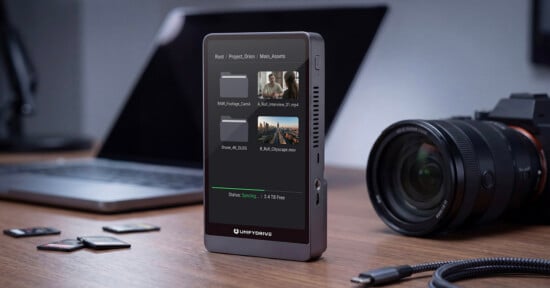 A portable external drive with a screen shows file folders and photo thumbnails, sitting on a desk near a laptop, a camera, SD cards, and a USB cable.