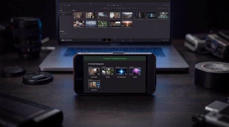 A smartphone displaying a media categorization screen sits on a dark desk in front of a blurred laptop showing video editing software; camera gear and tape are visible around them.