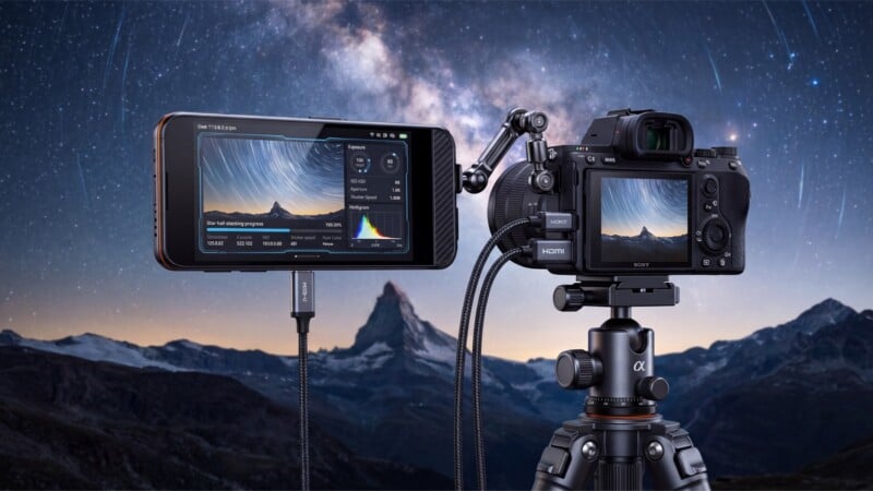 A digital camera on a tripod is connected to an external monitor displaying a long-exposure image of the night sky, with the Milky Way and mountains visible in the background.