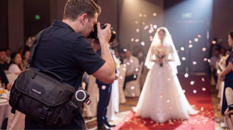 A photographer takes a picture of a bride walking down the aisle at a wedding ceremony, with flower petals in the air and guests seated on either side.