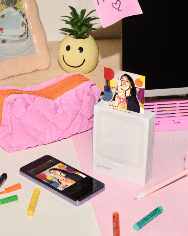 A workspace with colorful markers, a pink pencil case, a phone displaying a photo, a Polaroid printer printing the same photo, a smiley face plant pot, and a computer monitor with a pink sticky note.