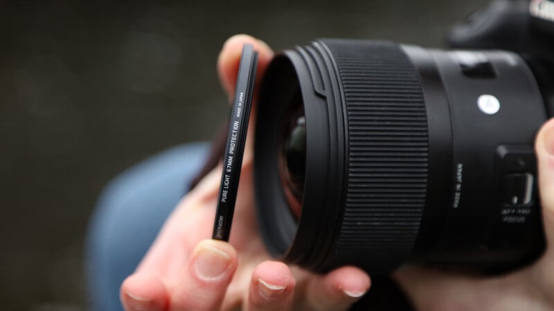 A close-up of a person’s hand holding a "Pure UV" camera lens filter near the front of a DSLR camera lens, preparing to attach it. The background is blurred.