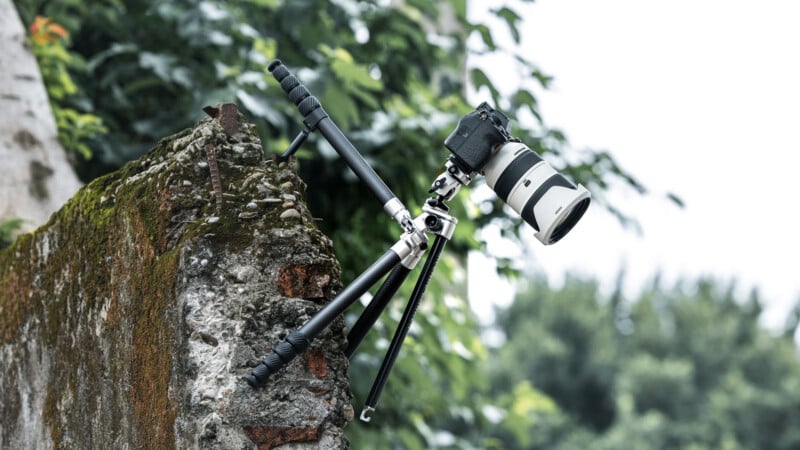 A camera mounted on a tripod is positioned at an angle on a rough, broken concrete wall, with green foliage and trees blurred in the background.