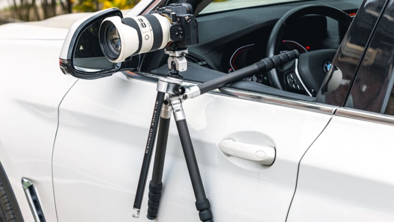 A camera with a large telephoto lens is mounted on a tripod that is set up through the open window of a white car, with the tripod legs resting inside and outside the car door.