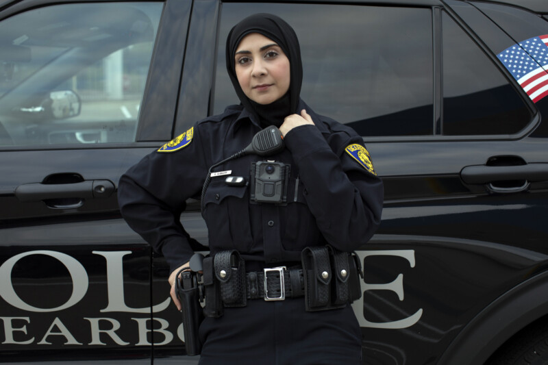 A police officer wearing a black uniform and a hijab stands in front of a police vehicle with an American flag visible. She looks confidently at the camera, with one hand resting on her duty belt.