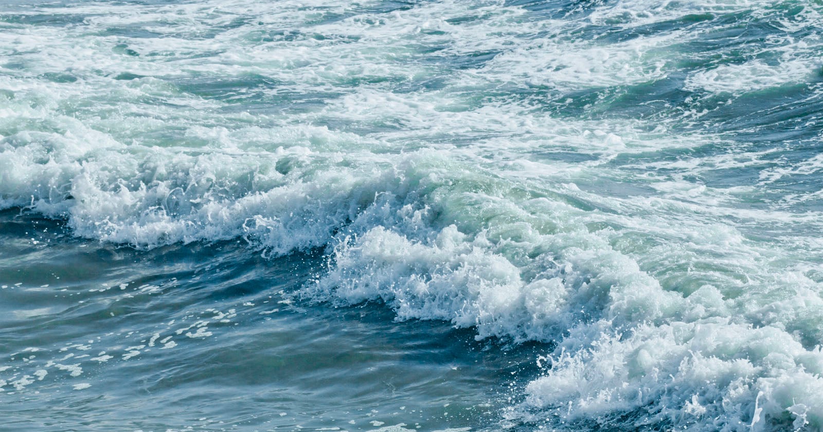 Waves crashing in the ocean, with white foam forming on the surface of the blue-green water under daylight.