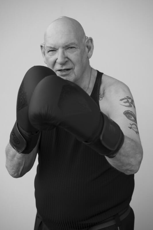 An older man with a bald head and tattoos poses with black boxing gloves raised in front of his face, wearing a sleeveless shirt and standing against a plain background.