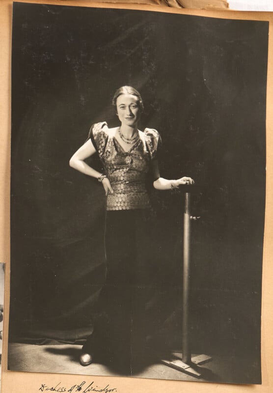 A woman stands indoors, wearing a shiny, short-sleeved top and dark pants, with one hand resting on a metal stand. The background is dark, and she faces the camera, smiling slightly.