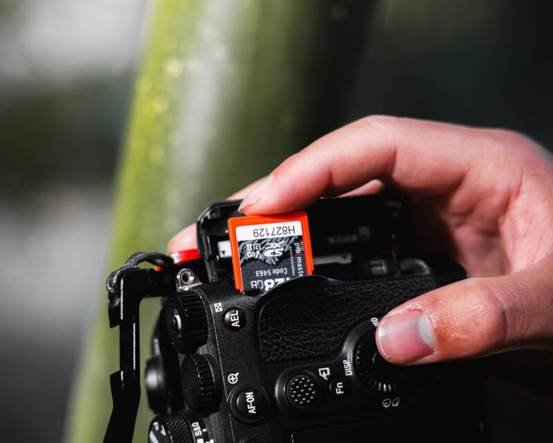 A hand inserts an orange memory card labeled "128 GB" into a black digital camera, with the background out of focus.
