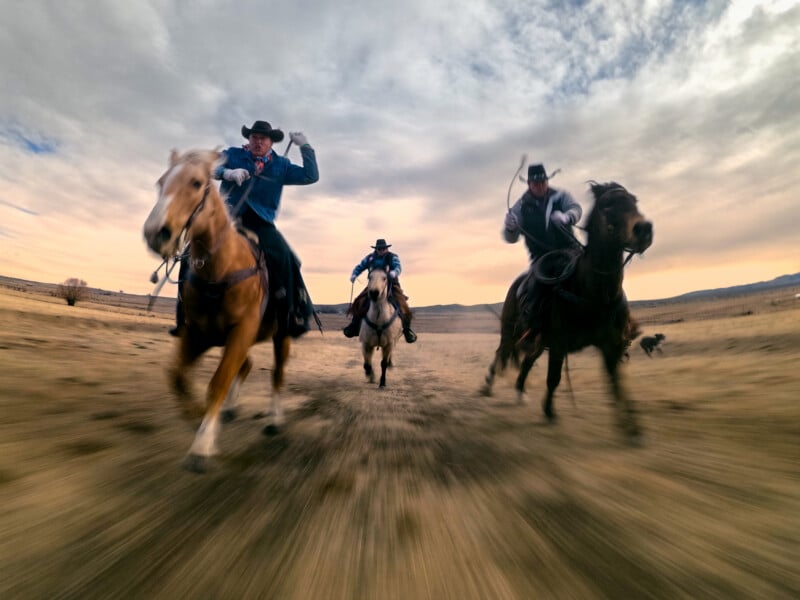 Three cowboys ride horses at full gallop across an open plain under a dramatic sky, with motion blur emphasizing their speed and energy.