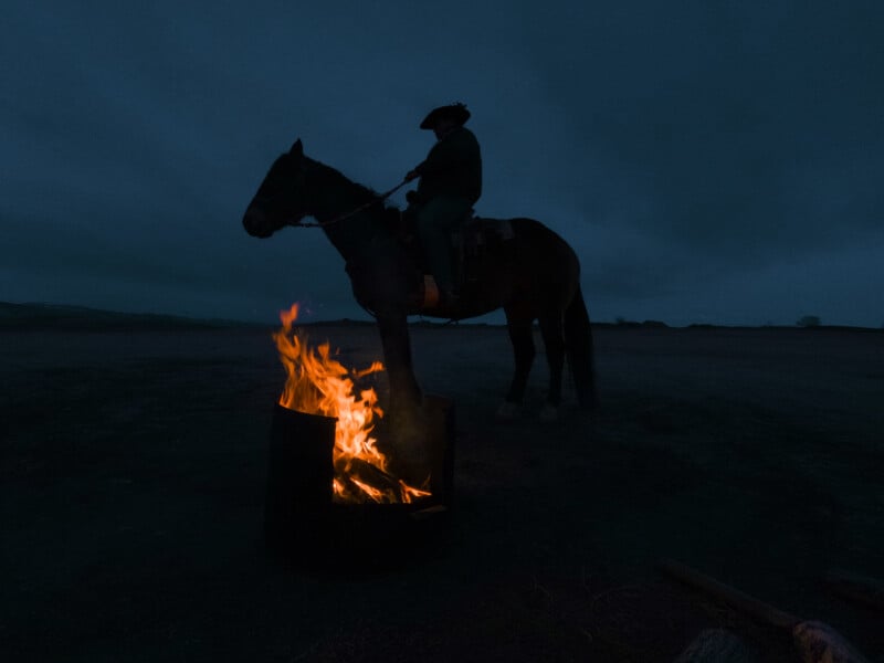 A person wearing a hat rides a horse near a blazing fire at night, silhouetted against a dark, cloudy sky on an open landscape.