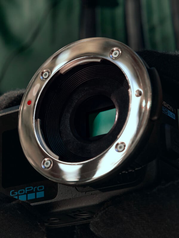 Close-up of a GoPro camera with its lens removed, revealing the sensor and metal lens mount. The camera is being held by a gloved hand, with visible screws and a red alignment dot on the mount.