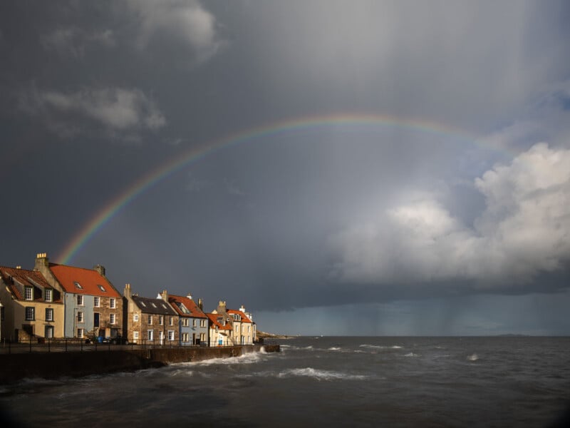A rainbow arcs across a cloudy sky above a row of seaside houses with red roofs, as waves crash against the shore under dramatic lighting.
