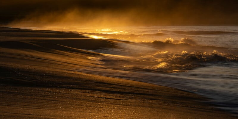 Golden sunlight shines on ocean waves crashing onto a sandy beach, creating mist and highlighting the textured sand at sunset or sunrise. The scene feels warm and tranquil, with dramatic light and shadows.
