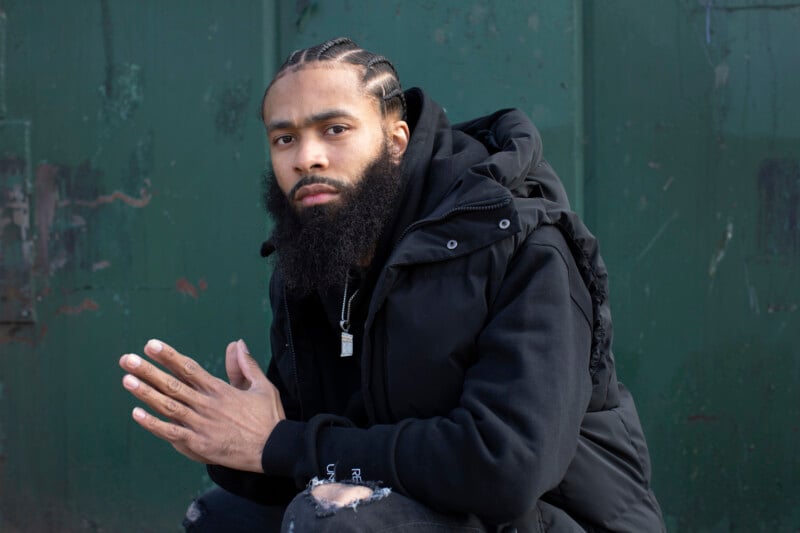 A man with braided hair and a full beard, wearing a black jacket and ripped black jeans, sits in front of a worn green wall, looking directly at the camera with his hands together.