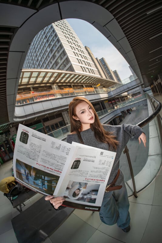 Una mujer se encuentra en el interior de un edificio moderno y lee una revista grande. Esta foto tiene un efecto de ojo de pez y muestra edificios altos, edificios curvos y una pasarela al aire libre al fondo.