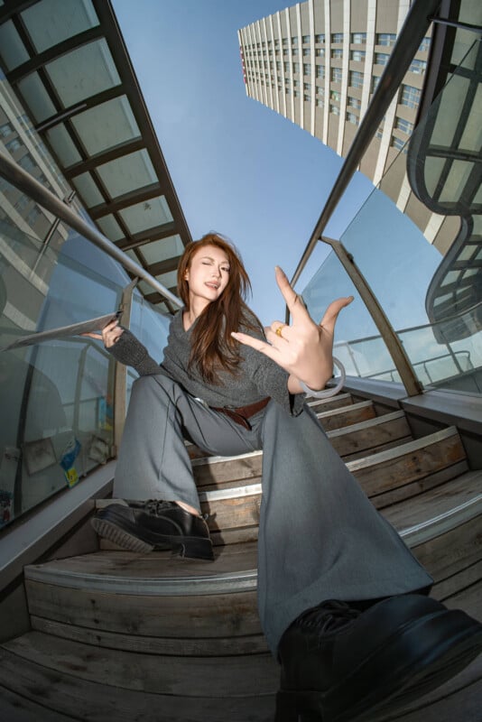 Una mujer joven con pantalones anchos y zapatillas de deporte posa con confianza en las escaleras al aire libre. Hace un gesto de rock 'n' roll hacia la cámara, con edificios modernos y cielos azules de fondo. Esta foto utiliza una lente gran angular.