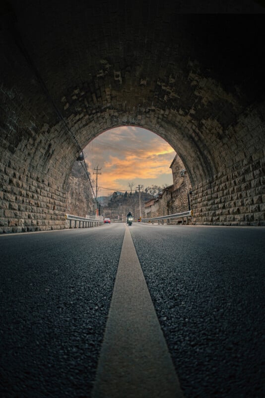 Un túnel de ladrillos de piedra conduce a la carretera al atardecer, y una línea central blanca conduce al horizonte. Un hombre conduce una motocicleta hacia el túnel, y afuera se pueden ver líneas eléctricas y edificios.