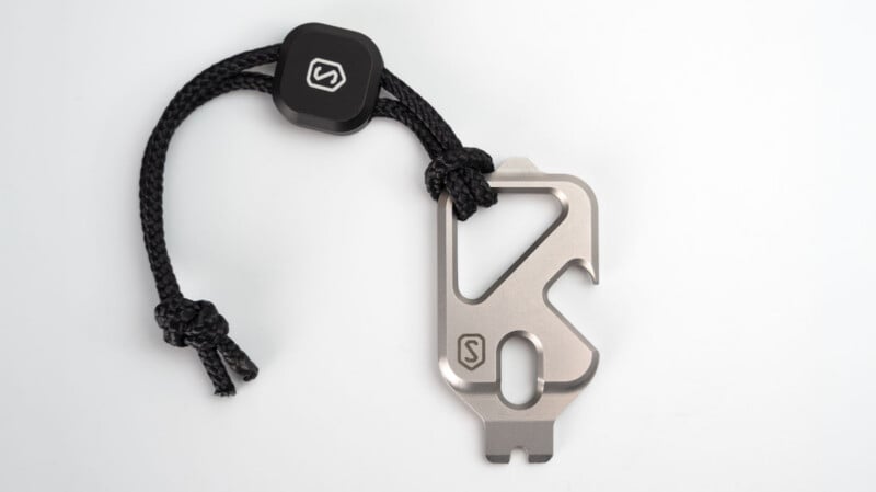 A silver multi-tool with a black braided cord and a logo-printed rubber tag, featuring various tool edges and cutouts, displayed on a white background.