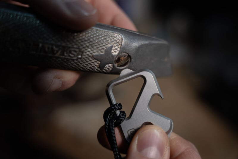 Close-up of hands holding a utility knife and a small metal multitool with a paracord loop, using the multitool’s hook to open or pry something from the utility knife.