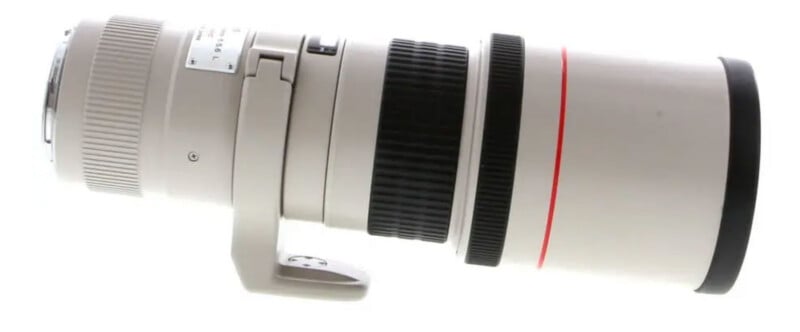 A white telephoto camera lens with a tripod mount collar, black grip rings, and a red ring near the front element, shown on a white background.