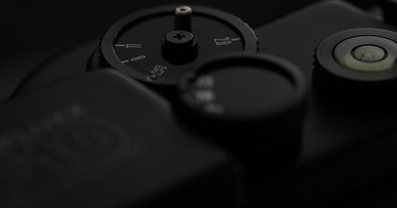 Close-up of a black camera with focus on its dials and buttons, including an aperture control dial and a bubble level, set against a dark background.