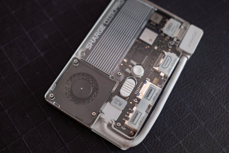 A close-up of a transparent electronic device showing internal components, including a cooling fan, labeled ports, circuit board, and visible branding that reads "SHARGE" on a dark textured surface.