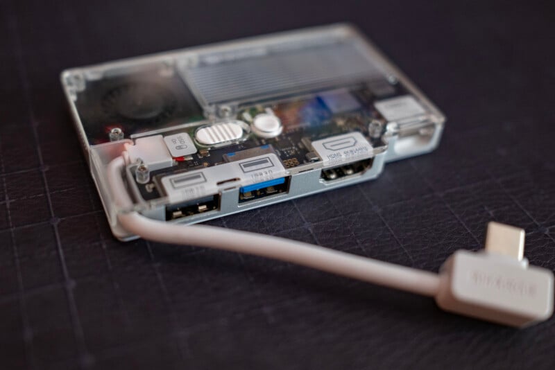 A close-up of a transparent electronic device case showing ports labeled USB, HDMI, and HDMI 2, with a white USB-C cable plugged into one of the slots, all set on a dark textured surface.
