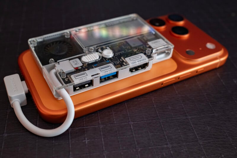 A transparent-cased mini computer with visible ports and a fan sits on top of an orange smartphone, both placed on a dark surface. A white USB-C cable is connected to the mini computer.