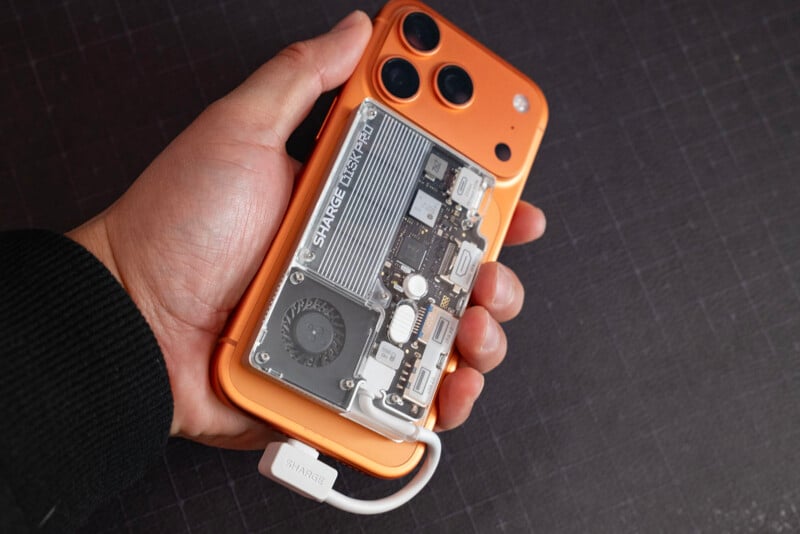 A hand holds an orange smartphone with a transparent case revealing internal components, including a visible fan and circuit board. A white cable labeled "STARPCIE" is connected to the phone.
