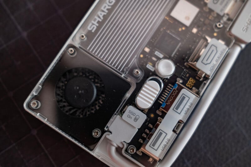 Close-up of a computer or electronic device’s internal components, including a cooling fan, circuit board, connectors, labeled ports, and visible microchips, all enclosed in a partially transparent casing.
