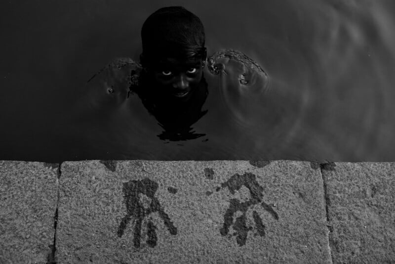 A child with only their head and shoulders above dark water looks up, eyes wide. Wet handprints are visible on the stone ledge in front of them. The image is in black and white.