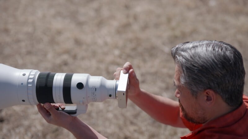 A man with gray hair is adjusting a large white telephoto camera lens outdoors, with dry grass visible in the background.