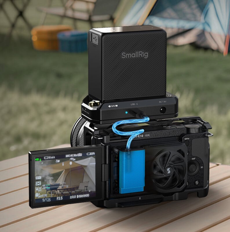 A digital camera with an external battery pack labeled "SmallRig" attached on top, positioned on a wooden surface outdoors with a tent and lawn chairs in the blurred background. The camera screen shows a live view of the scene.